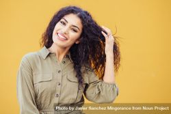 Female in army green jumpsuit smiling in front of mustard wall playing with hair 5z18n0