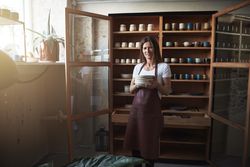Smiling woman in leather apron holding a stack of handmade plates in front of cabinet in her shop