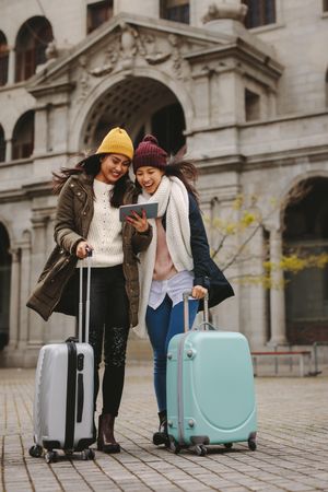 Two young women with suitcases smiling at tablet