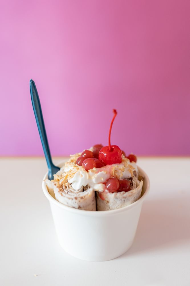 Close up of a rolled ice cream decorated with strawberry bubbles, nut toppings and a cherry on top
