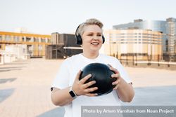 Smiling blonde woman with medicine ball, smart watch and headphones on a rooftop 5ryeZ0