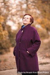 Older woman in purple coat standing near autumn trees 0WLWM5