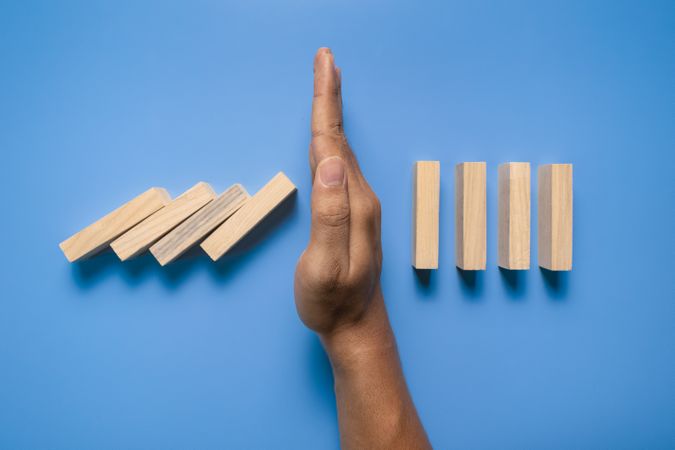 male hand stop falling wooden blocks isolated on blue background. domino effect or risk protection strategy concept