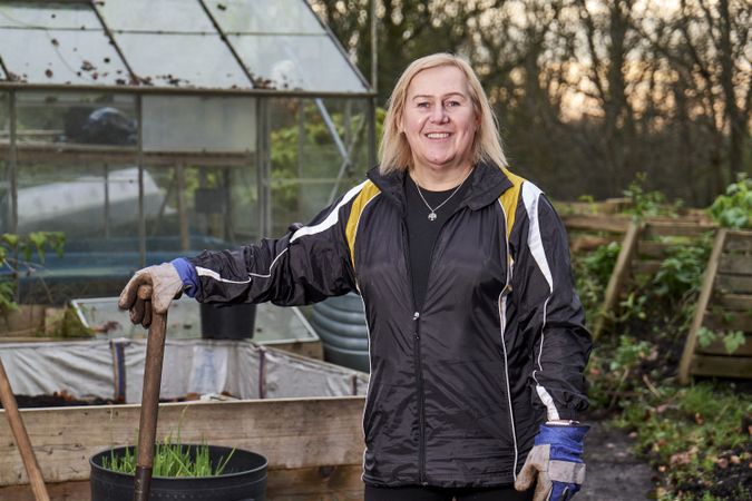 Older woman smiling outside greenhouse with shovel