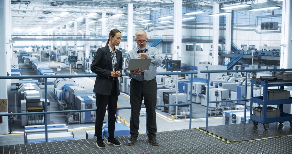 Mature Engineer and Young Female Technician Having a Conversation and Using Laptop Computer. Specialists Monitoring, Analyzing System Reports at a Modern Electronics Factory