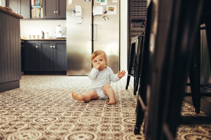 Cute baby sitting on the kitchen floor with hand in mouth