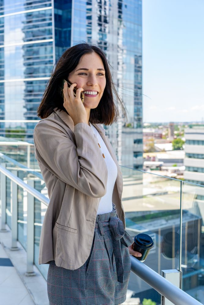 A woman on her office balcony talks on her cell phone and holds a paper cup of coffee during her break.