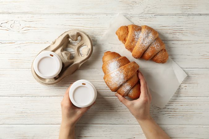 Female hand holding paper cup of coffee and croissant on wooden table, top view