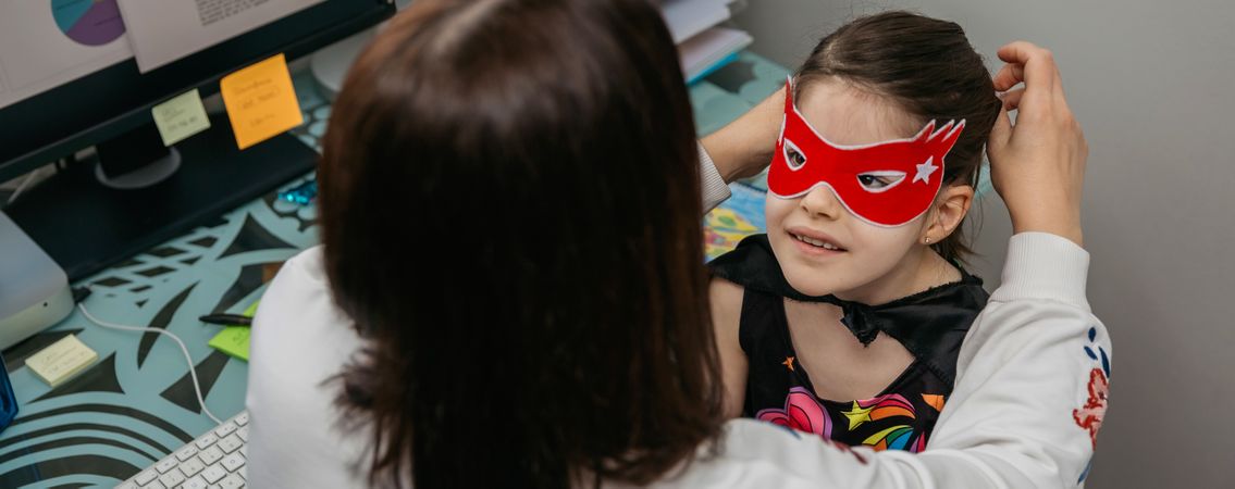 Woman teleworking and putting costume mask on her daughter