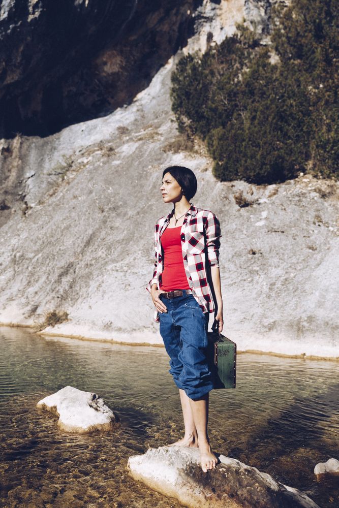 woman standing on rock with case near clear water in lake