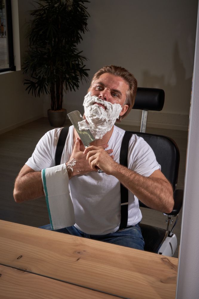 Adult barber shaving with knife in front of mirror