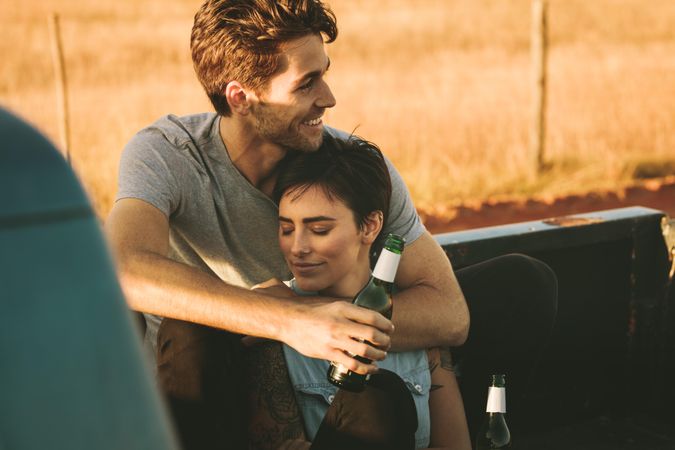 Man and woman holding each other while enjoying a bottled beer during road trip