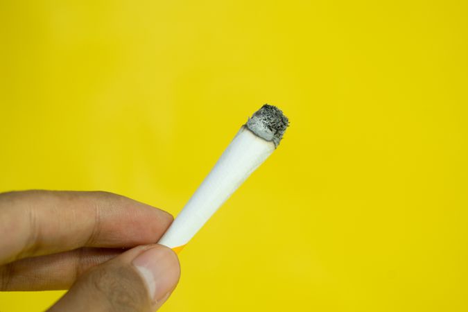 Side view of hand holding lit hand rolled cigarette on yellow background