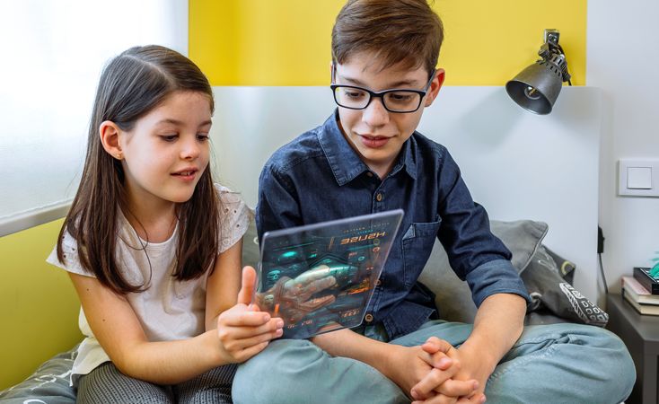Happy children playing games and having fun with futuristic transparent tablet sitting over bed.