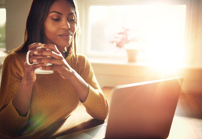 Woman holds coffee mug as she works on laptop in sunny room