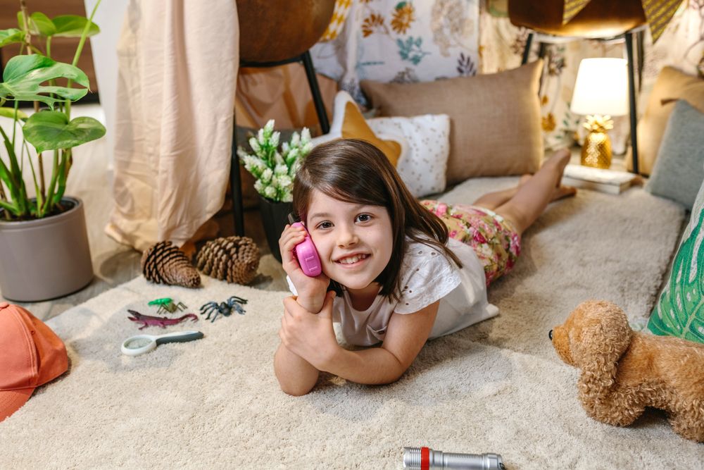 Smiling little girl looking at camera while using walkie-talkie lying over rug surrounded by cushion