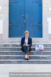 Smiling female in blazer sitting in front of blue door outside with coffee bxXnrb