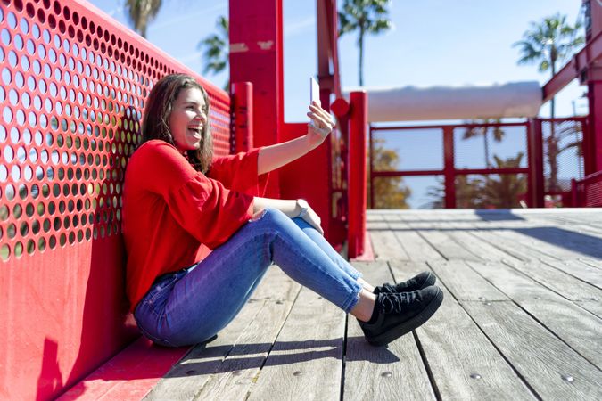 Side view of smiling young woman in red sitting taking selfie on mobile phone outdoors on a bright day