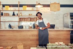 Portrait of happy young man wearing an apron and hat at a cafe counter holding a cup of coffee 5kReA0