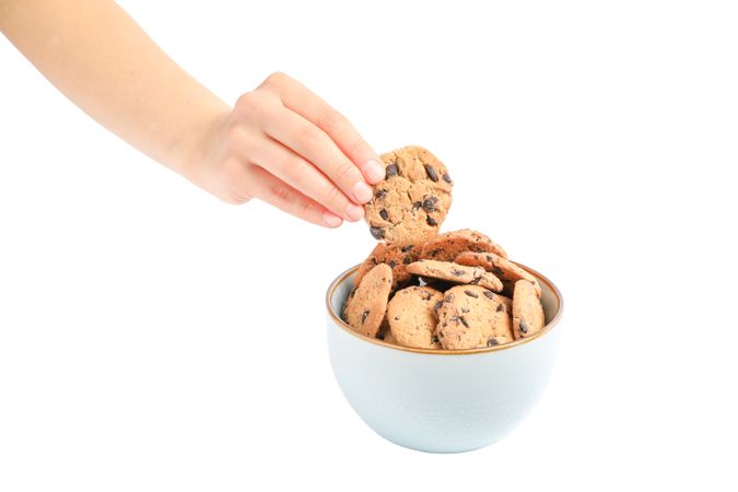 Young woman holding tasty chocolate chip cookie over bowl on studio background, closeup