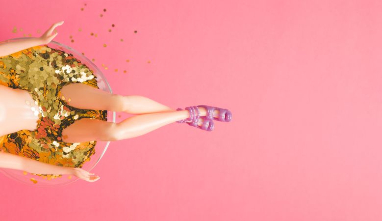 Top view of doll bathing in martini glass full of gold glitter on pink background