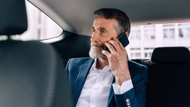 Mature man with grey beard on phone in backseat of car