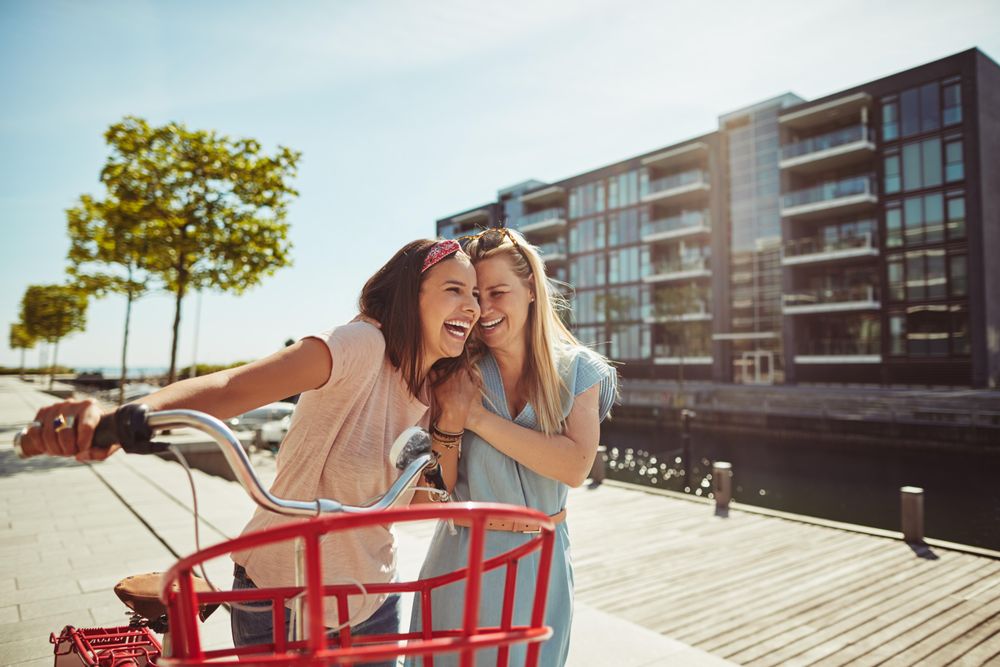 Two happy female friends laughing with arms on shoulders while walking with bike, copy space