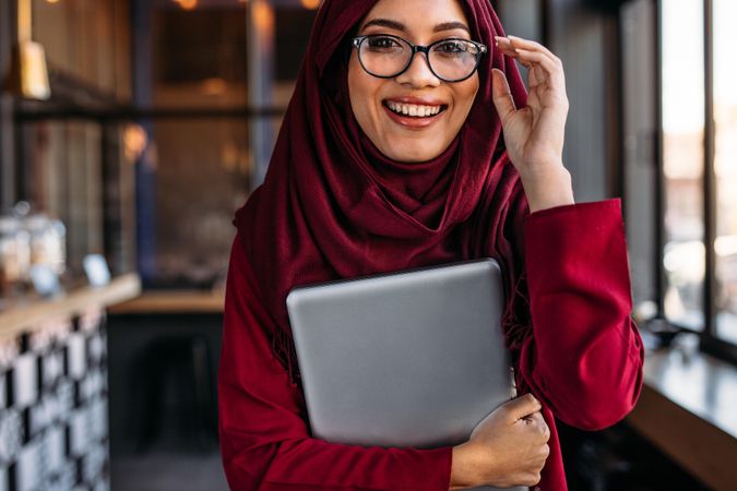 Muslim woman with laptop in hand adjusting her eyeglasses at coffee shop