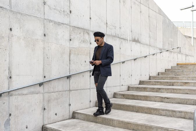Young Latino gay man wearing a fashionable hat and sunglasses, walking down the stairway while texting on his smartphone