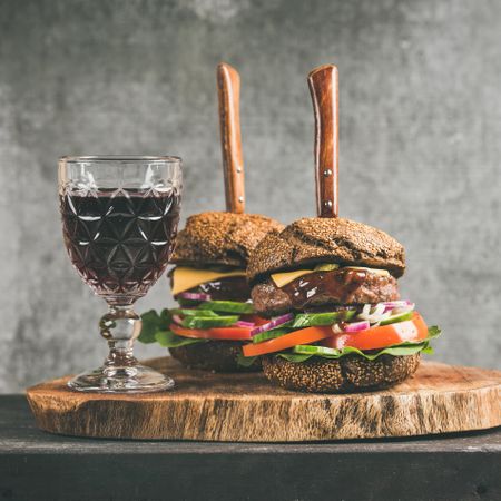 Two cheeseburgers, stacked with fresh vegetables, on wooden board, with wine, knifes, square crop
