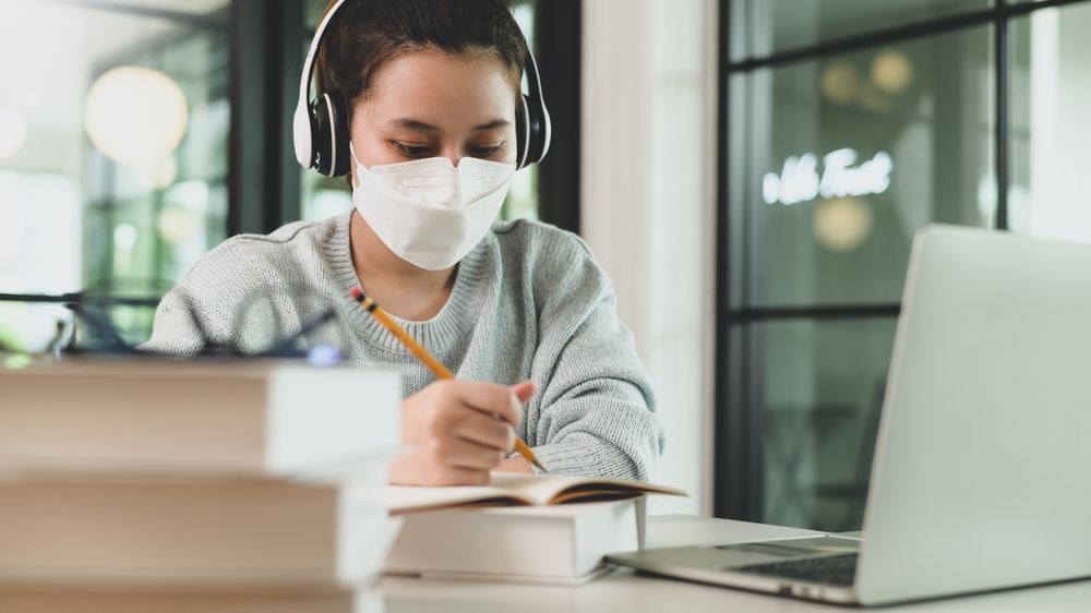 Asian woman with stethoscope wearing medical mask taking notes and studying online with laptop.