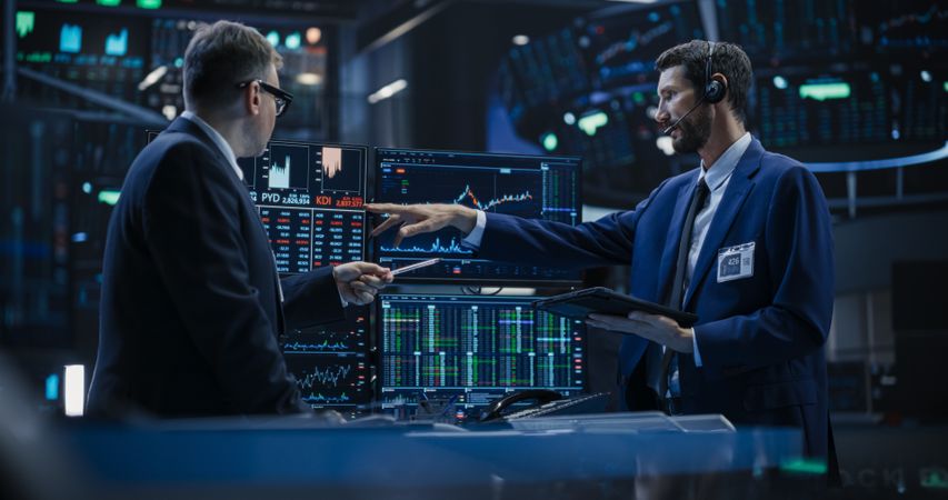 Team of Two Handsome Stock Exchange Brokers Using Tablet Computer, Chatting About the Asset Management Strategy for an International Business Client, Working in a Office Building with Computer Screens