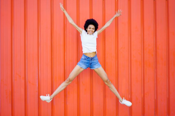 Woman in shorts and t-shirt jumping for joy against outdoor red wall