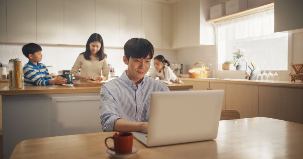 Portrait of Young Korean Father Using Laptop To Work from Home While his Wife and Kids are Working Together to Prepare Lunch.