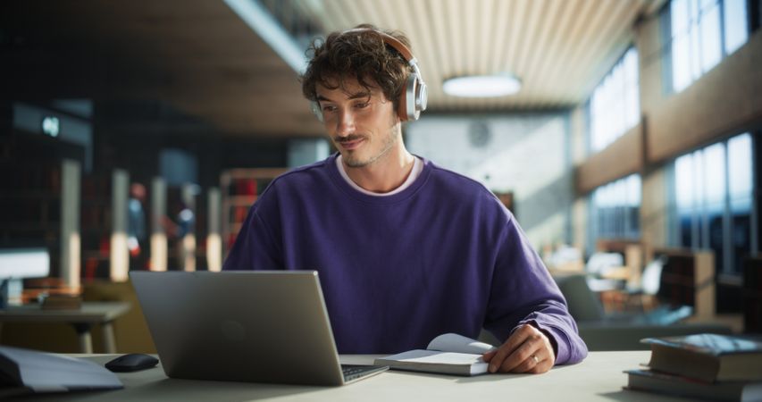 Student Writing an Essay on Laptop Computer, Thinking About the Topic, Brainstorming, Finding Solutions. Young Stylish Man Studying in a Public Library, Listening to Lecture in Headphones