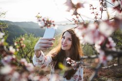 Young happy woman taking selfie on smartphone near trees on hill