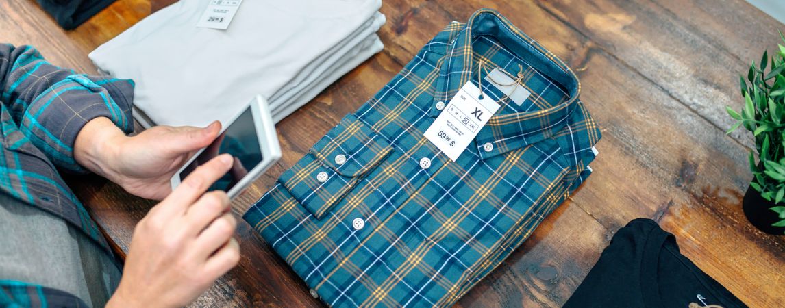 Banner of unrecognizable female entrepreneur checking stock on phone next to apparel stacks on store
