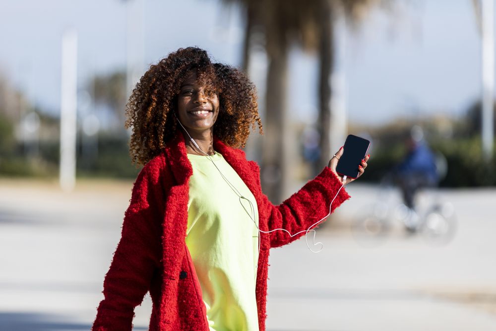 Confident happy female in colorful clothes dancing to music on phone on street with palm trees