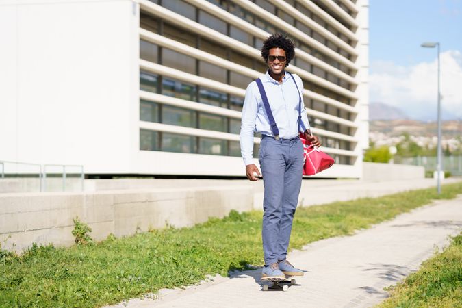 Smiling man in sunglasses  with afro hair riding skateboard next to office