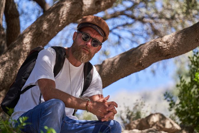Unemotional bearded male traveler sitting on tree branch