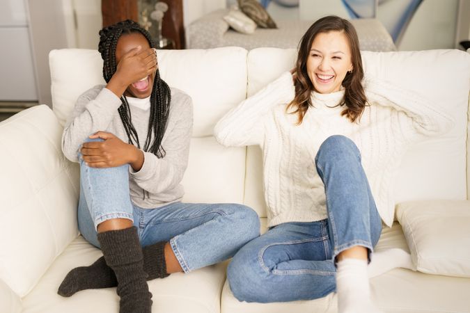 Two women in jeans and sweater joking together on a sofa
