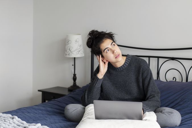 Young female thinking while sitting on her bed with a laptop