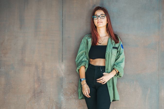 Stylish confident bright woman in glasses leaning on wall and looking at camera