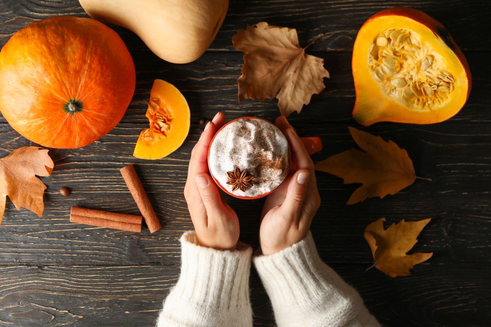 Female hands hold cup of pumpkin latte on wooden background with pumpkin, cinnamon and leaves