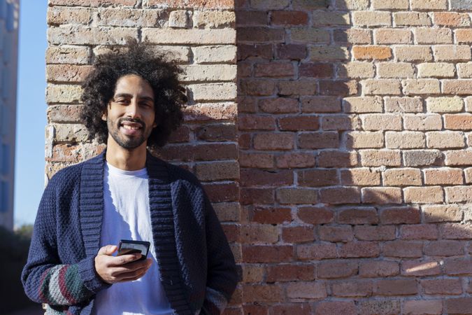 Smiling Black man holding his smartphone while leaning on a brick wall outdoors on sunny day