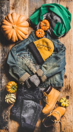 Flat-lay of vintage denim jacket, sweater, jeans, woolen scarf, hat, yellow boots, glasses, pumpkin,