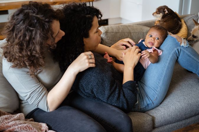 Two new moms snuggling on sofa holding their baby lovingly with dog