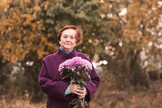 Older woman in purple coat holding flower bouquet standing near yellow trees