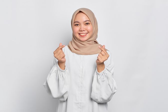 Muslim woman in headscarf and light blouse and smiling and making Korean heart shape with fingers
