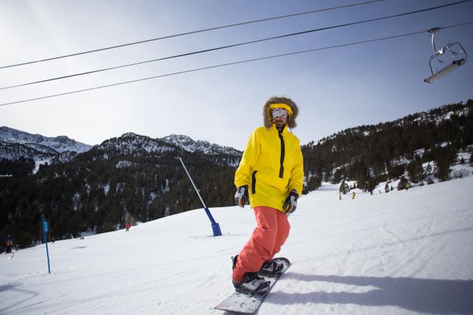 Man in yellow parka and goggles with snowboard with ski lift in background